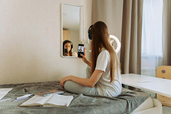A beautiful girl teenage with long hair ,generation Z in headphones at the mirror in room takes selfie on smartphone, admires herself. Home life, lifestyle. Selective focus