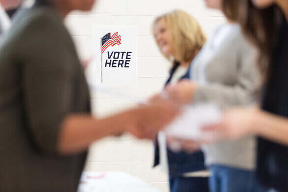 Citizens line up at a polling place, encouraged by a 'Vote Here' sign and American flag.