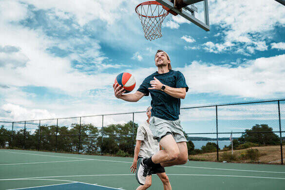 An man does a reverse layup while playing one-on-one with an older friend at an outdoor basketball court in Portland, Maine with waterfront views.
